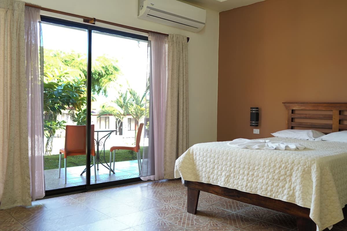 A hotel room with a bed, a wooden headboard, a sliding glass door leading to an outdoor area with a table and chairs, and a wall-mounted air conditioning unit.