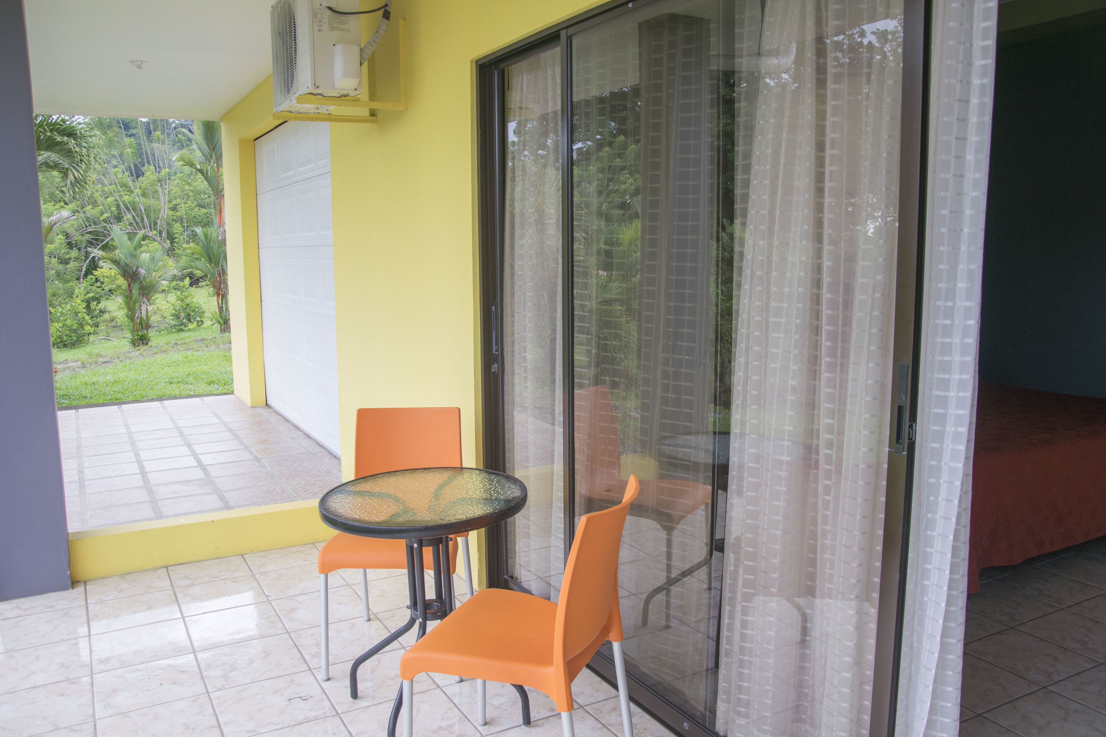 A room with a glass door leading to a balcony, a small table with two chairs, and a bed with a red cover.
