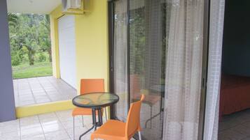 A room with a glass door leading to a balcony, a small table with two chairs, and a bed with a red cover.