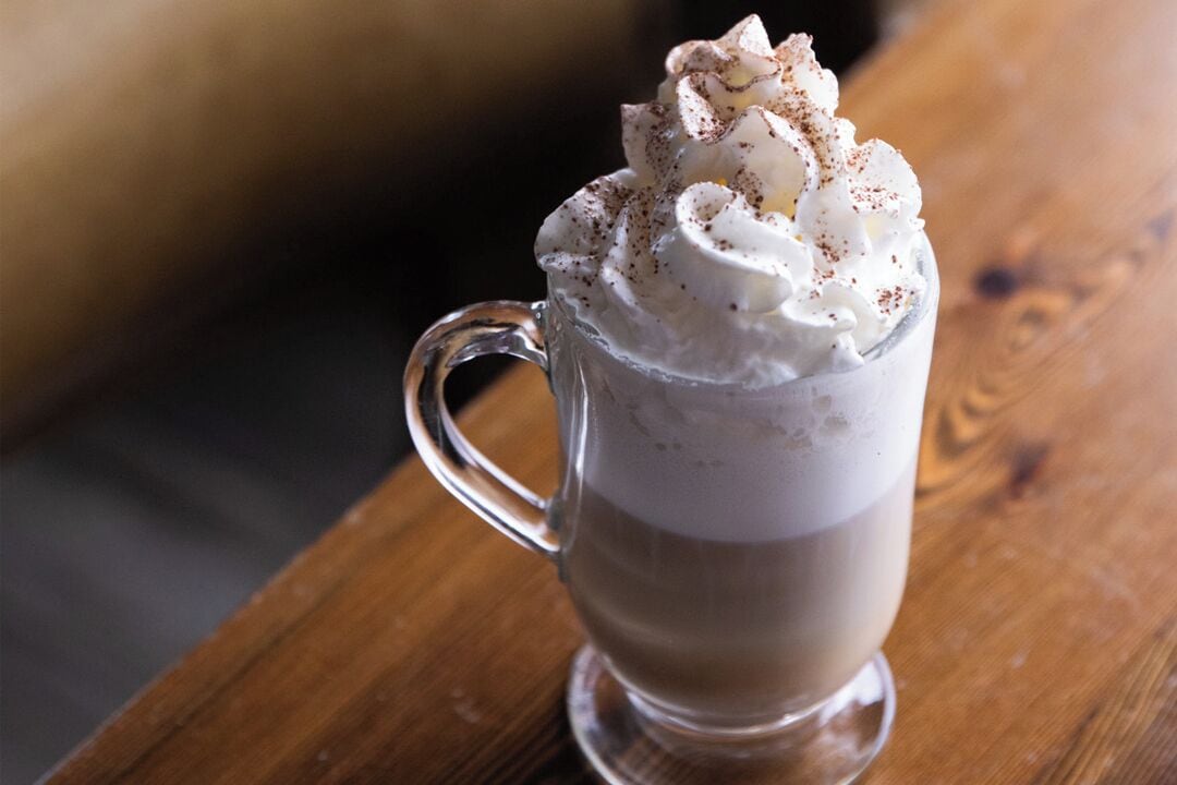A clear glass mug with a handle, filled with a layered beverage and topped with whipped cream and a sprinkle of cocoa powder.