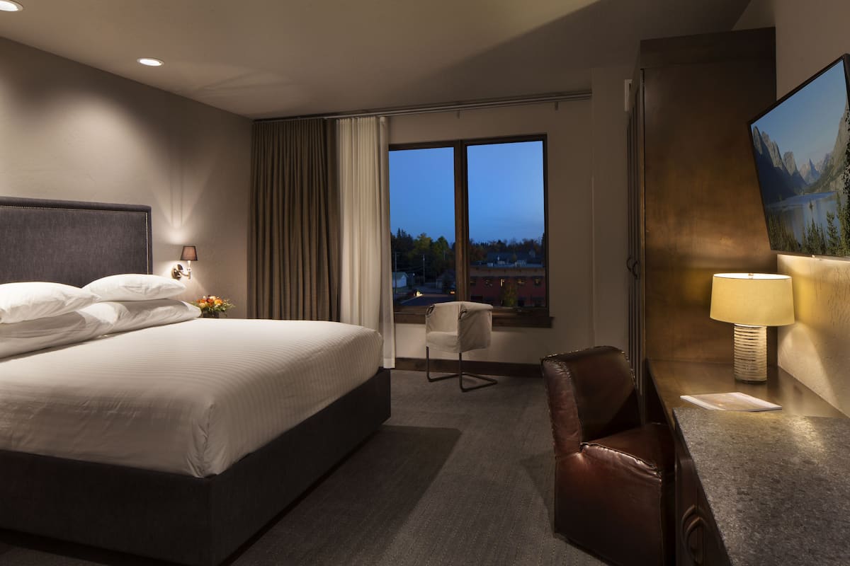 A hotel room with a large bed, a desk with a lamp, a chair, and a view of the mountains through the window.