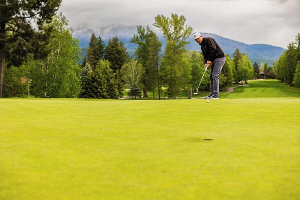 A golf course with a golfer in mid-swing, surrounded by trees and mountains in the background.