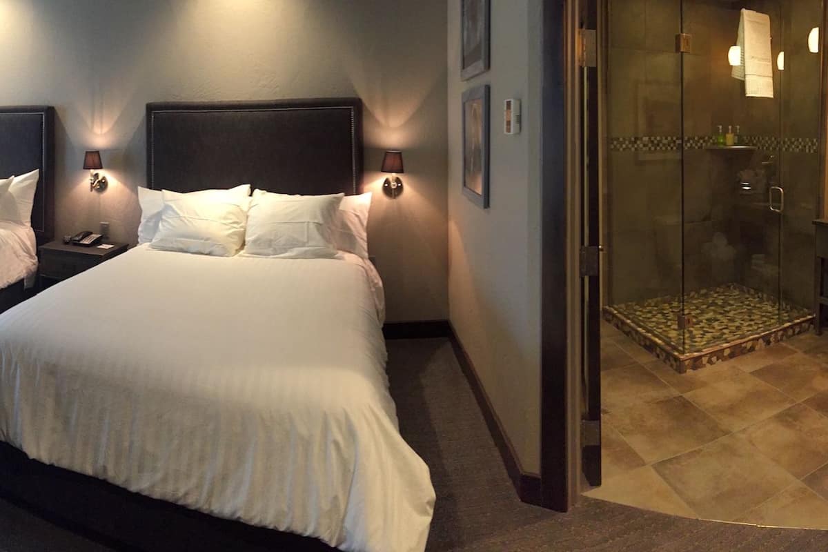 A hotel room with two beds, bedside lamps, a glass shower enclosure, and a small table with toiletries.