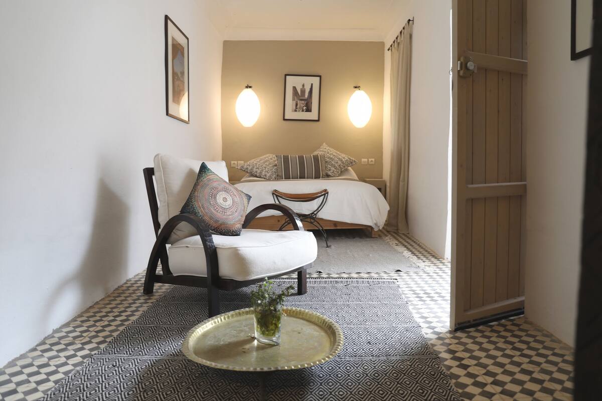 A hotel room with a checkered floor, a bed with pillows, a chair, a small table with a vase, and wall-mounted lights.