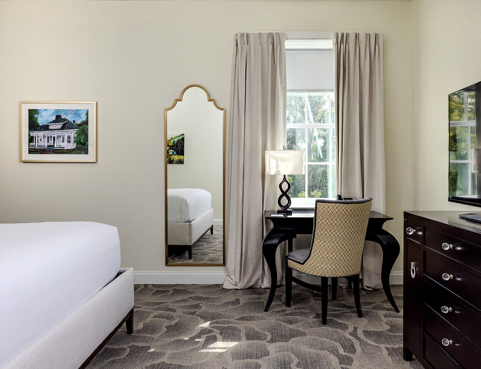 Quarto de hotel com cama, escrivaninha com luminária, cadeira, cômoda, espelho e janela com cortinas.