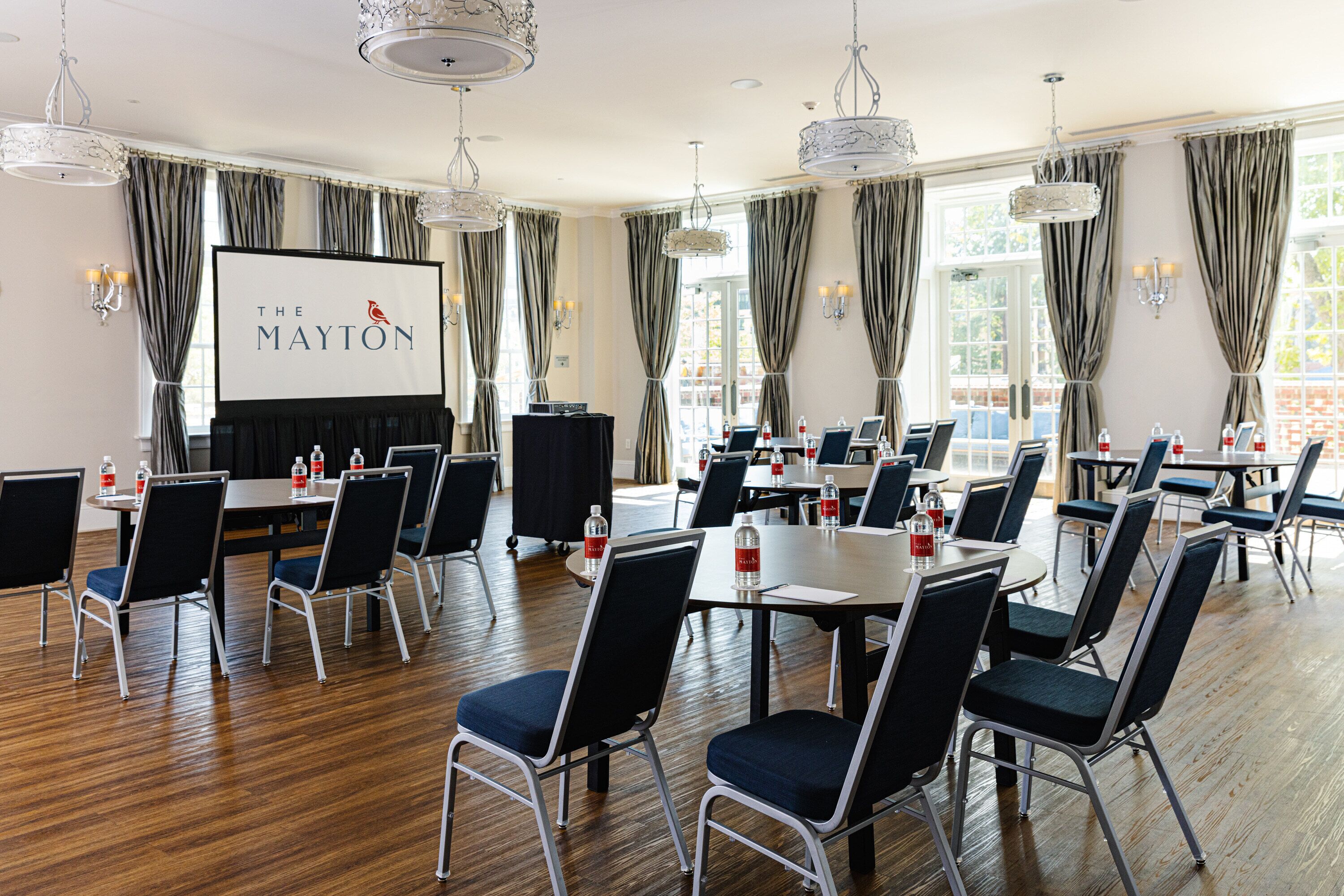 A conference room with multiple tables and chairs, a projector screen with "The Mayton" logo, and large windows with curtains.