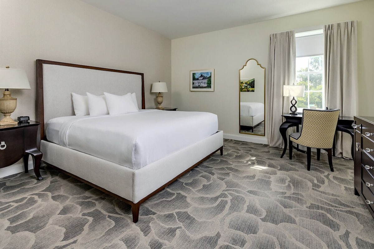 A hotel room with a large bed, a desk, a chair, a mirror, and a dresser.