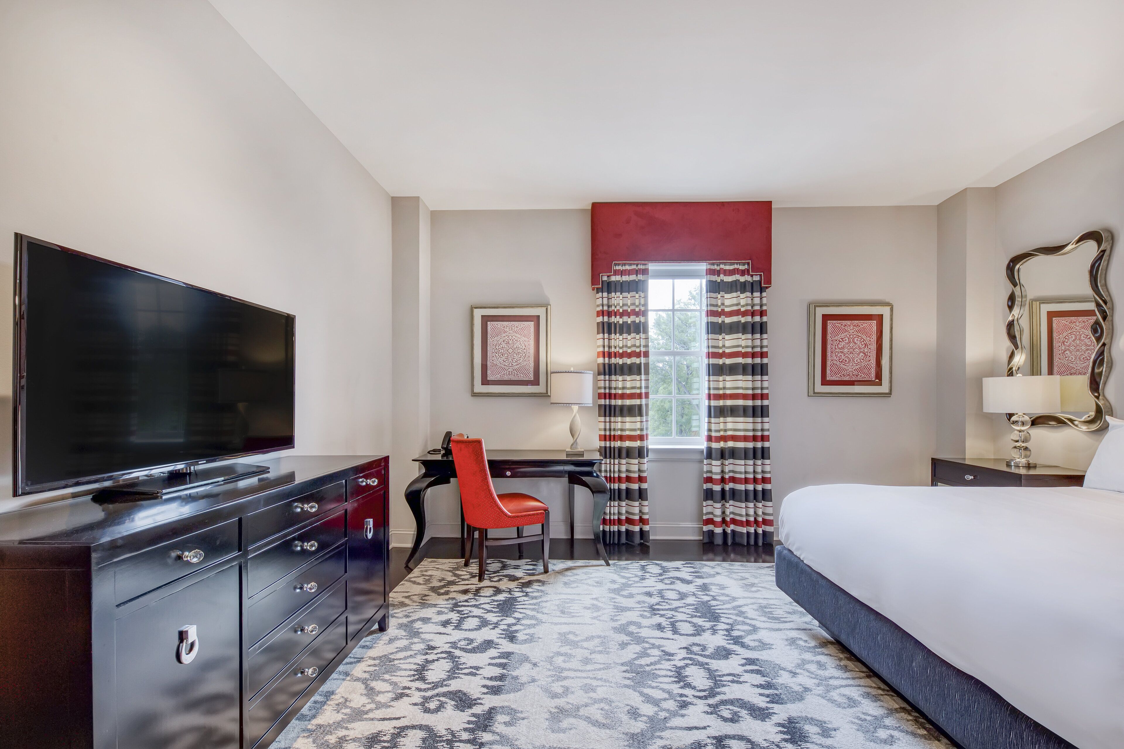 A hotel room with a large flat-screen TV, a desk with a chair, a bed, and a window with striped curtains.