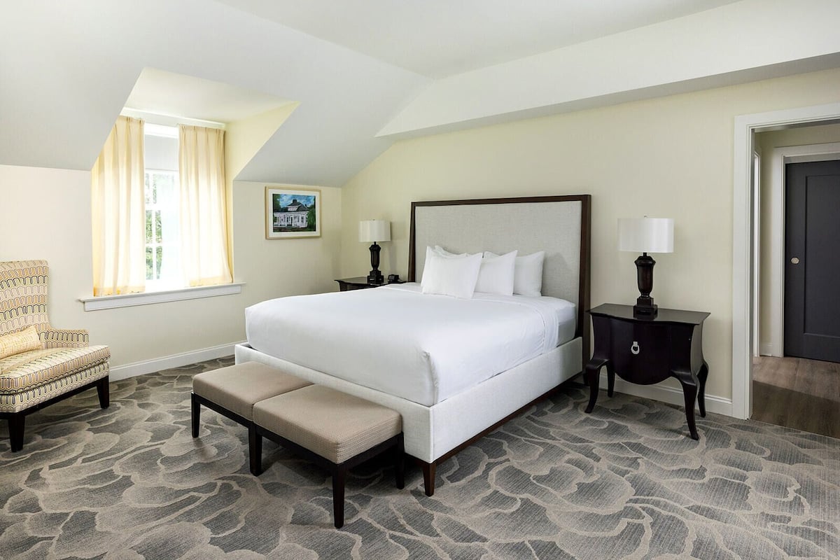 A hotel room with a large bed, a bench, a chair, a nightstand, a lamp, and a door.