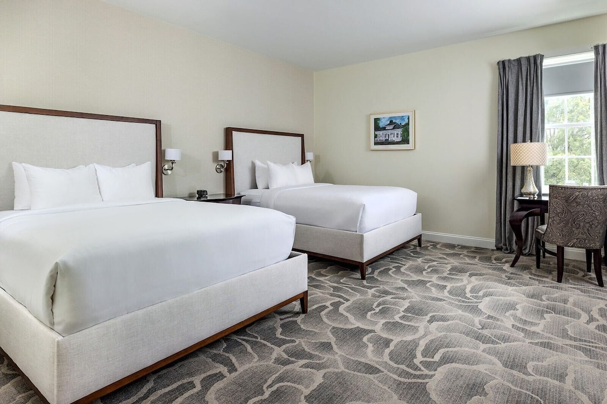 A hotel room with two beds, a desk, a chair, a window with curtains, and a painting on the wall.