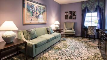 A room with a green sofa, two side tables with lamps, a patterned rug, a chair, and a large painting on the wall.