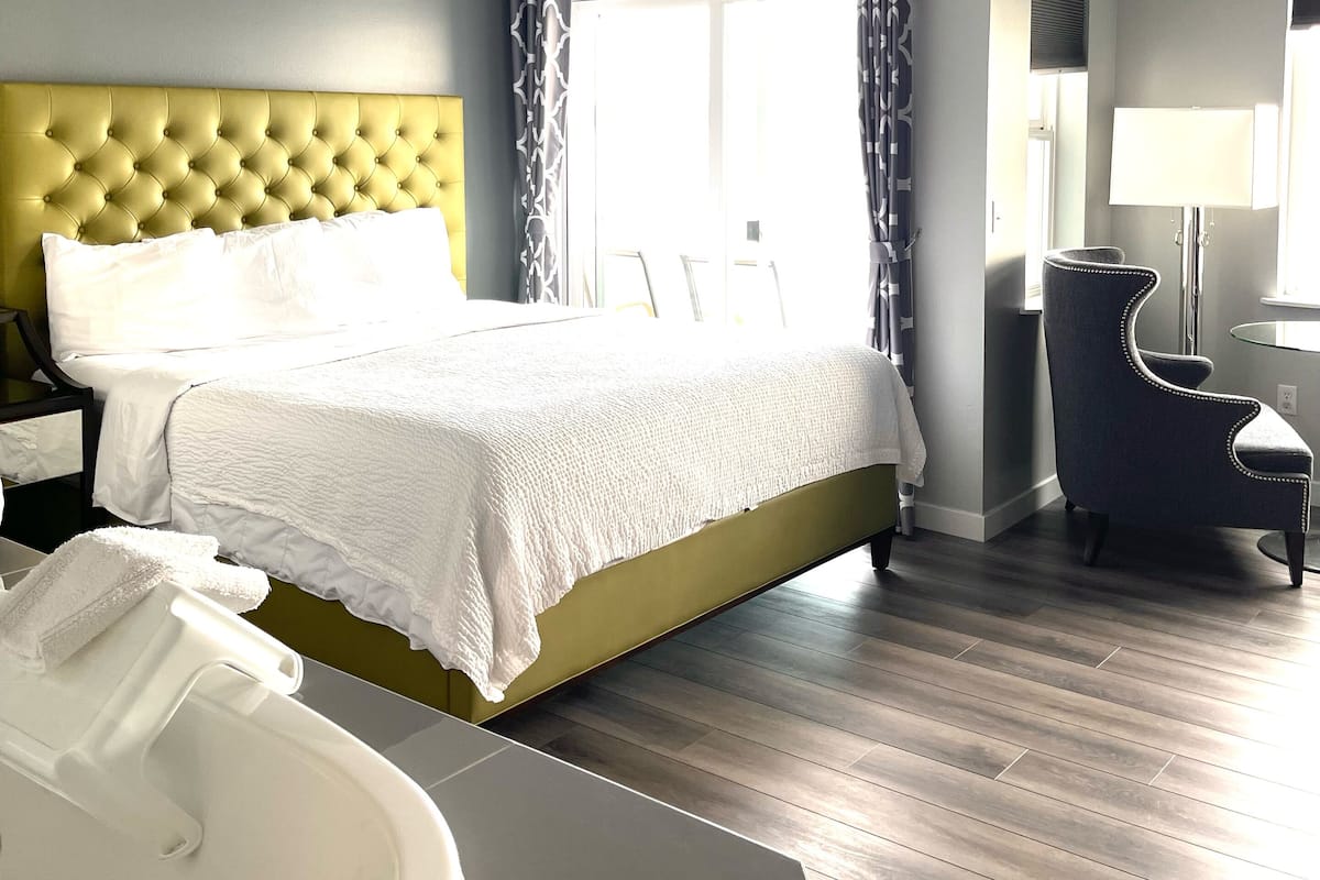 Una habitación de hotel moderna con una cama grande, una zona de estar con una silla, un escritorio y un ventanal amplio.