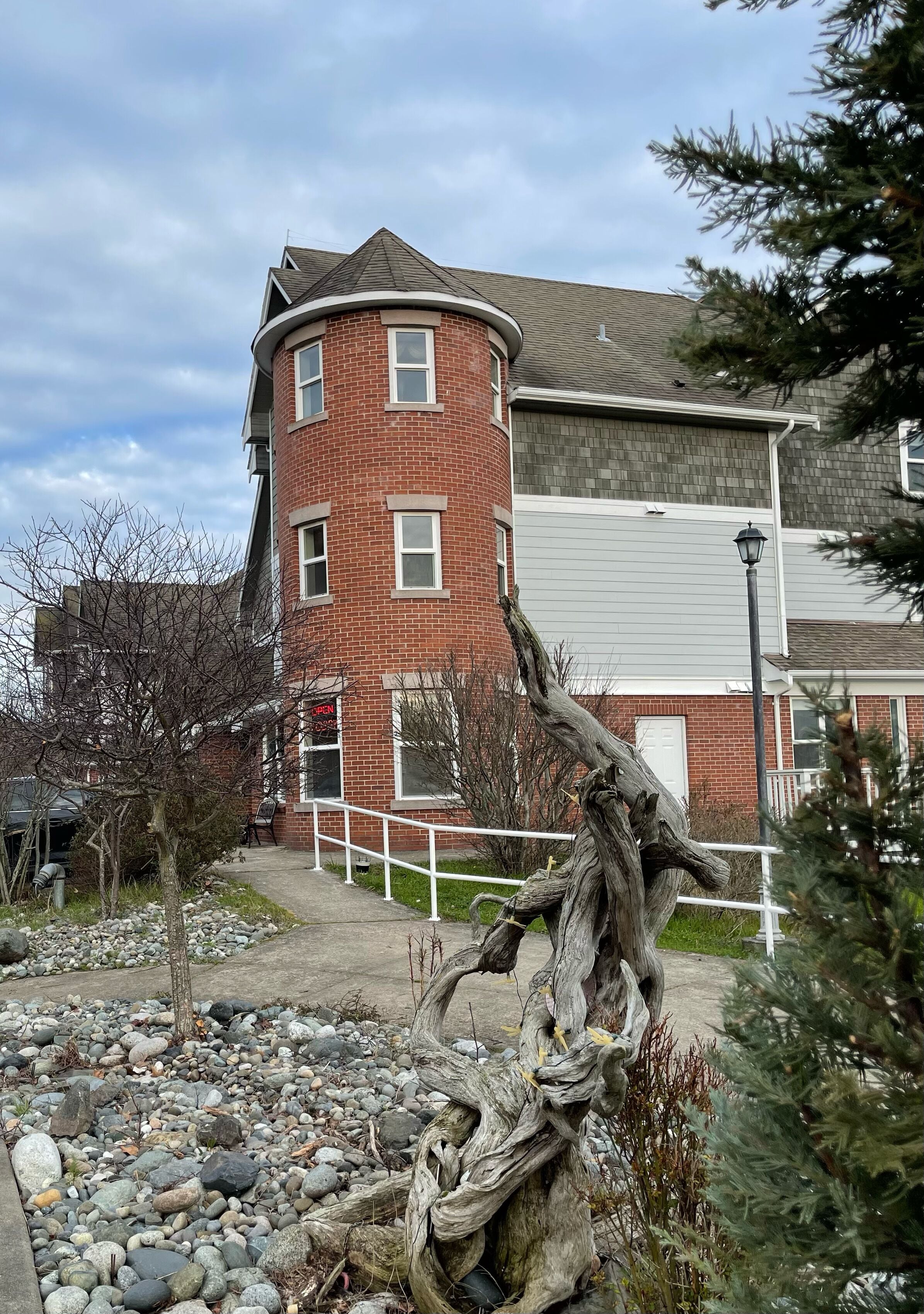A brick building with a curved corner, a white railing, and a large tree in the foreground.