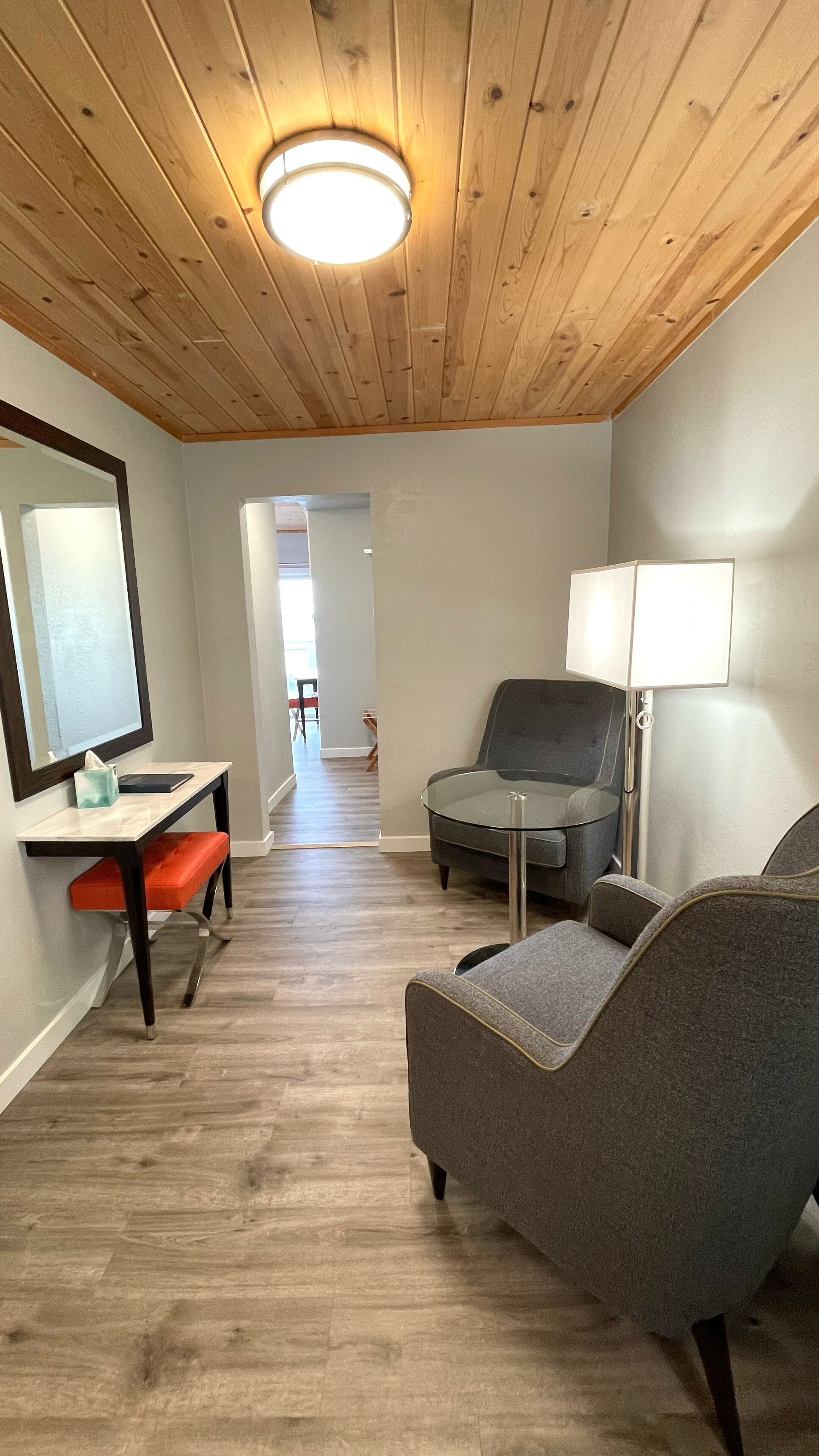 Une chambre moderne avec un plafond en bois, un luminaire rond au plafond, un fauteuil gris, une petite table avec un miroir et une lampe.