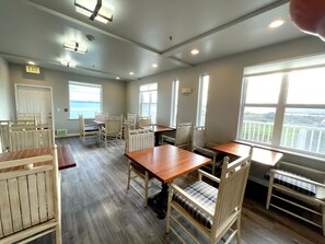 A dining area with wooden tables and chairs, a view of the sea through large windows, and an exit sign on the wall.