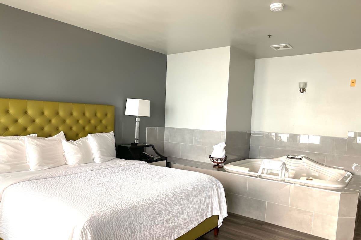 Habitación de hotel con una cama grande, una mesita de noche con lámpara, una pequeña mesa con teléfono, bañera y espejo.