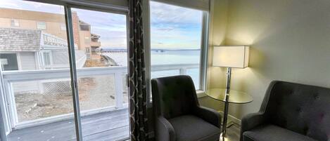 A room with two armchairs, a small round table, and a floor lamp. Large windows offer a view of a building and a body of water.