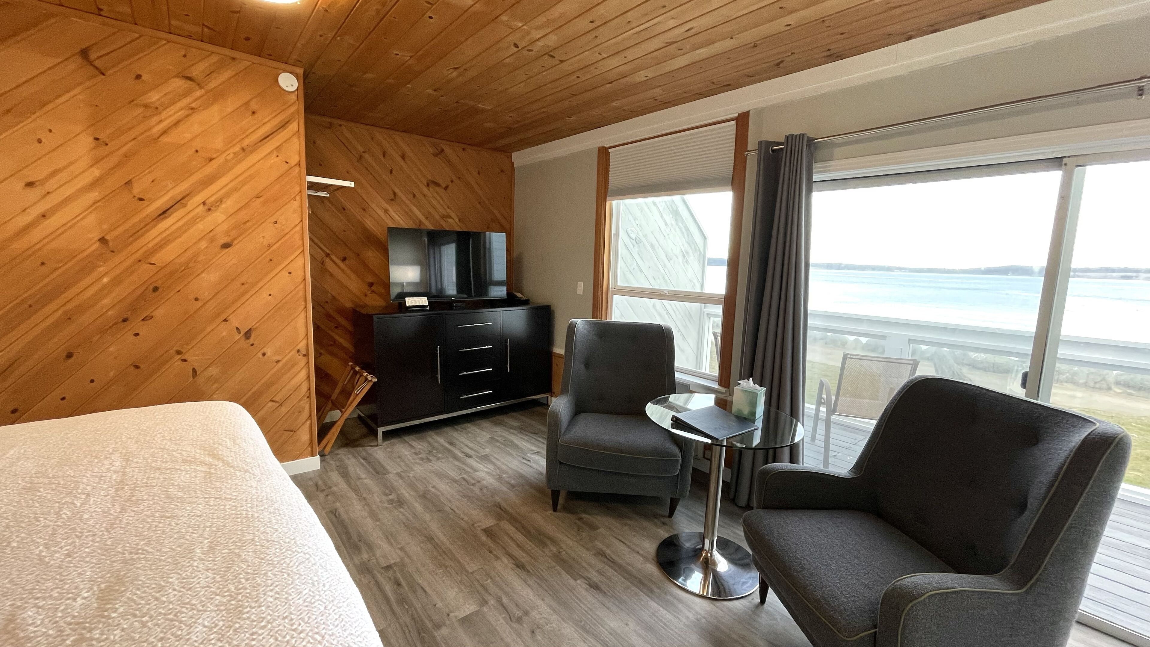 A hotel room with a wooden ceiling, a bed, a TV on a black cabinet, a small table with a chair, and a large window with a view of the sea.
