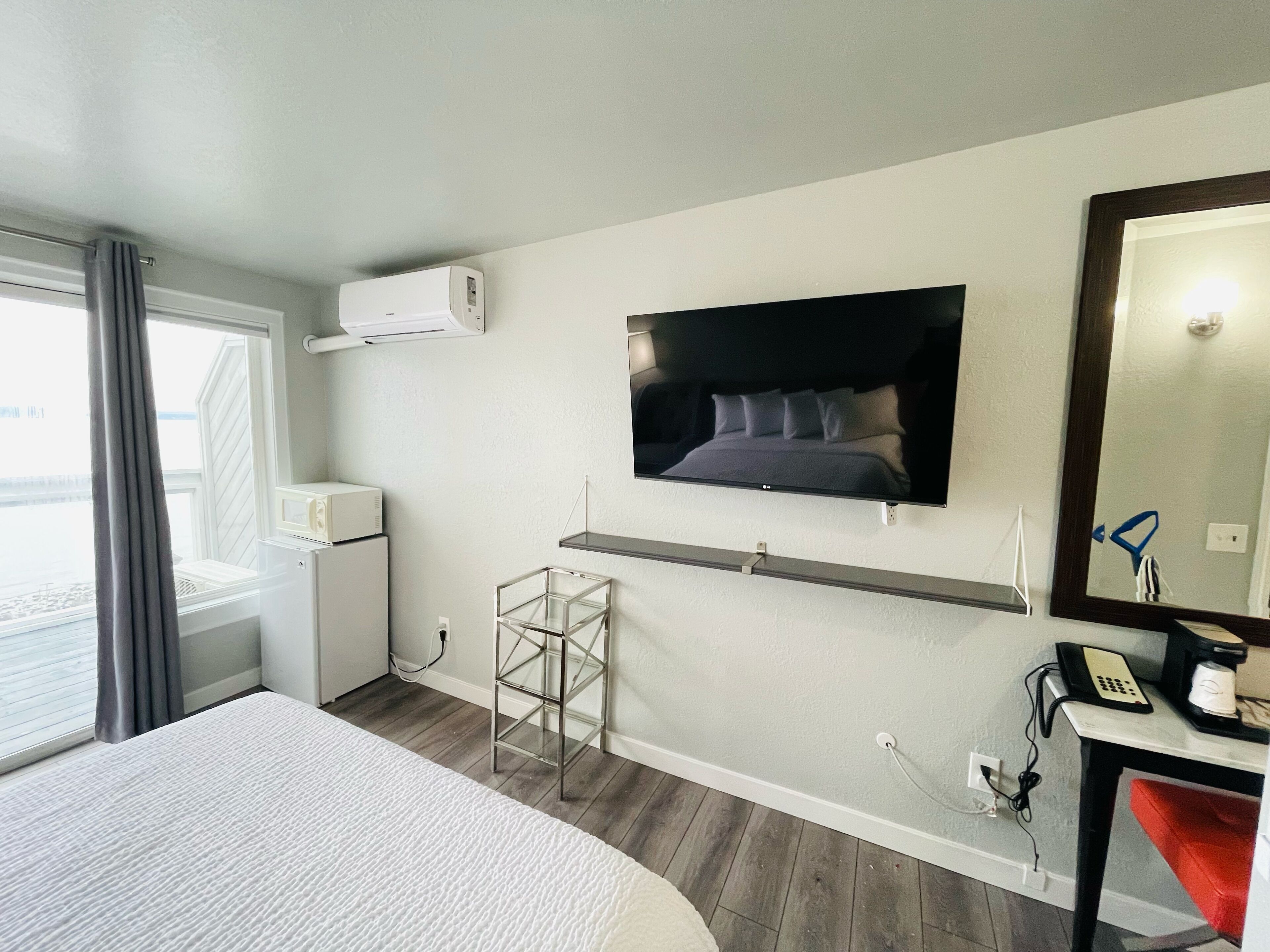 Habitación de hotel con cama, televisor montado en la pared, escritorio con teléfono y cafetera, y ventana con cortinas.