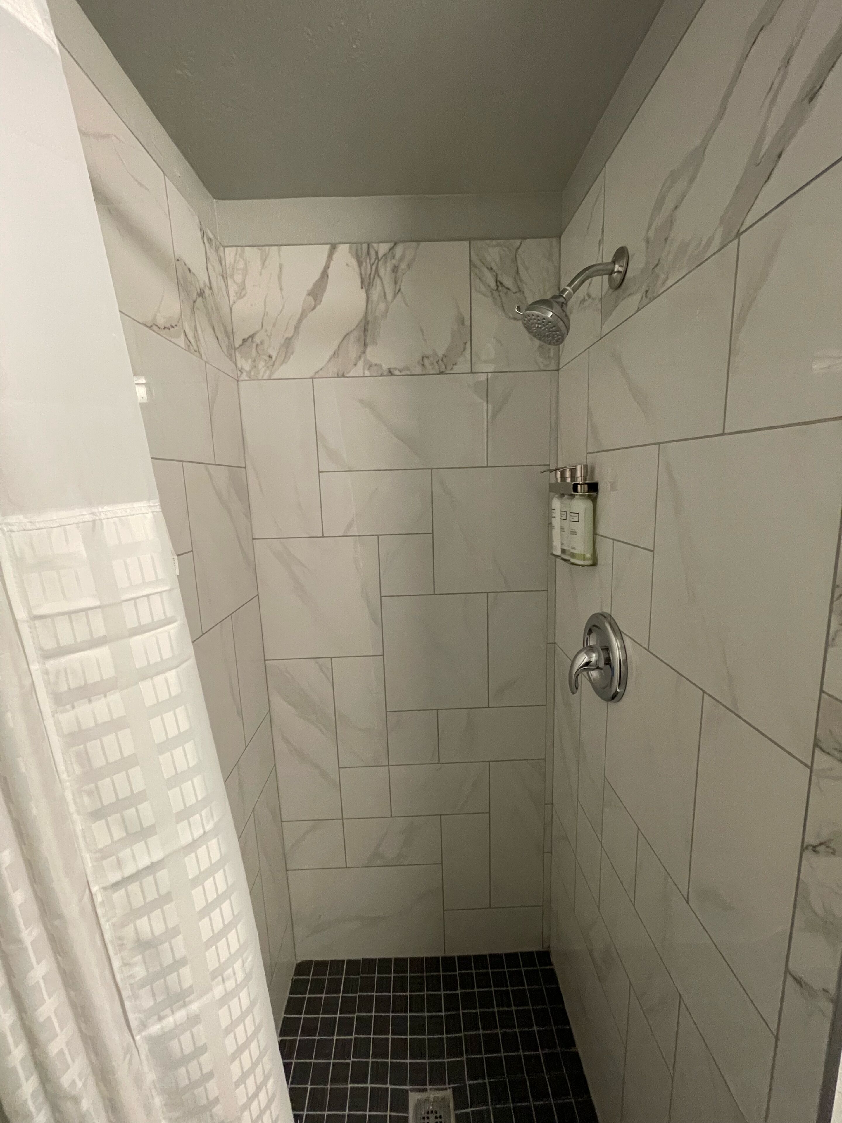 Ducha con azulejos blancos y de mármol, un cabezal de ducha y una botella de champú.