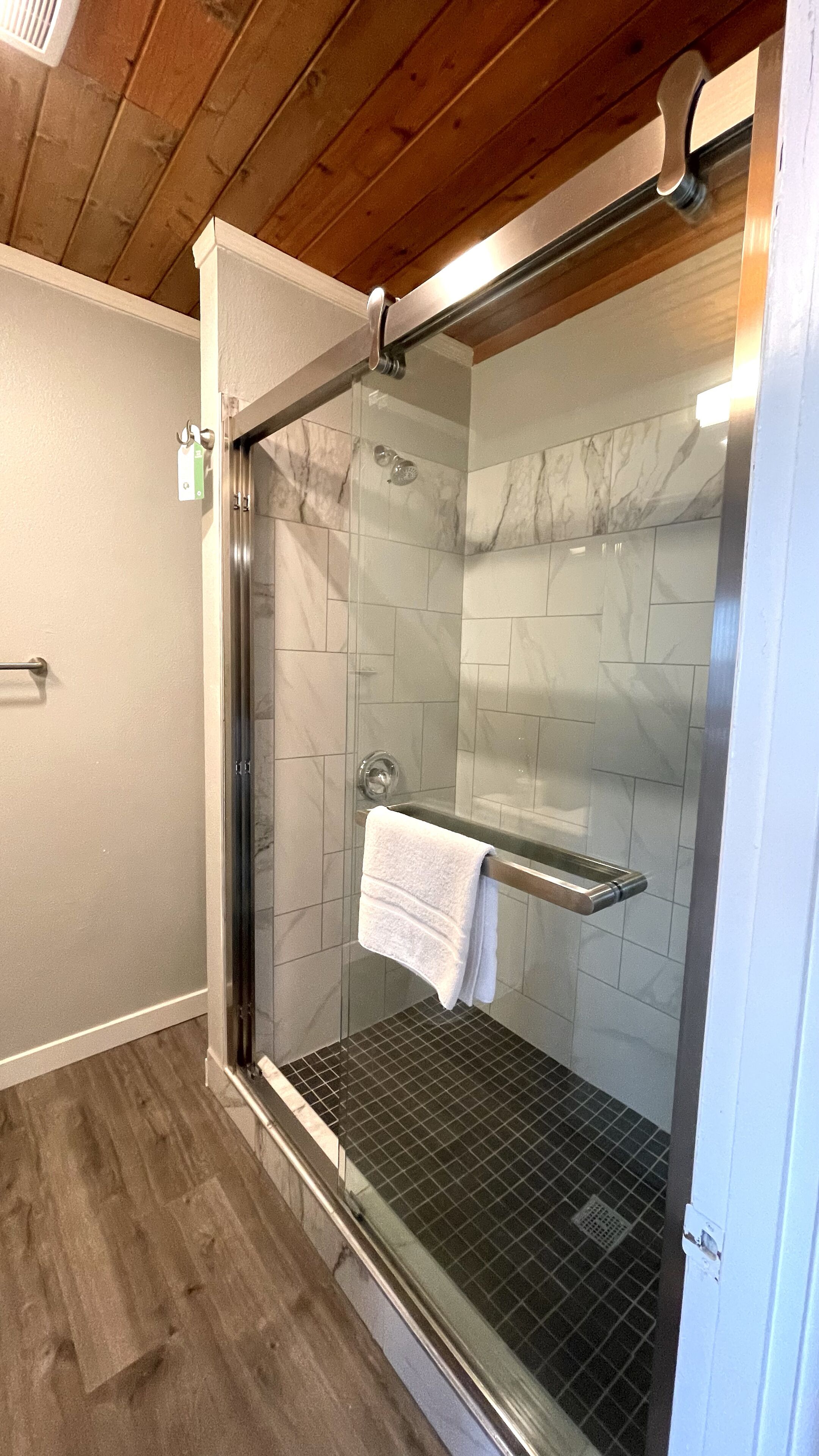 A glass-enclosed shower with a white towel hanging on a rack, tiled walls, and a wooden ceiling.