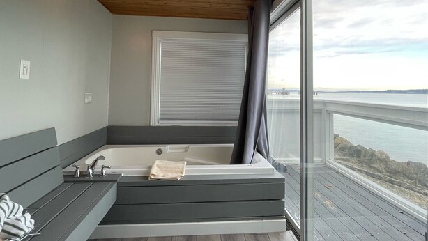 A modern bathroom with a bathtub, a bench, and a large window offering a view of the water. - Tides Inn & Suites (Port Townsend)