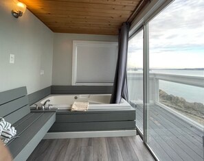 A modern bathroom with a bathtub, a bench, and a large window offering a view of the water. - Tides Inn & Suites (Port Townsend)