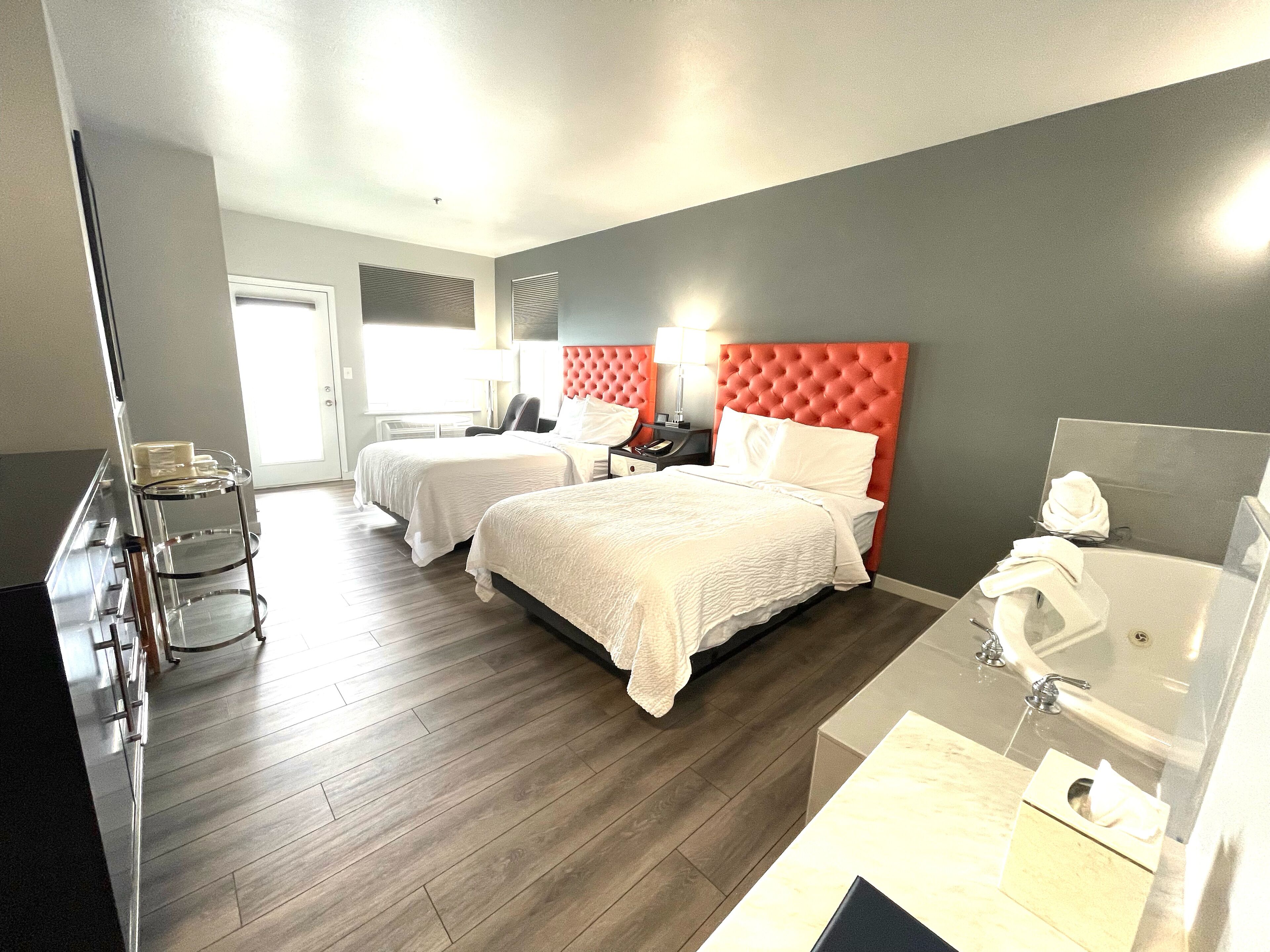 Habitación de hotel con dos camas, un escritorio, una silla, un tocador con espejo y un baño con lavamanos e inodoro.