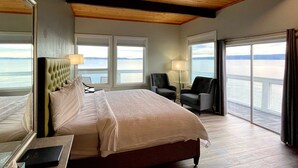A hotel room with a large bed, two armchairs, a desk, and a view of the water. - Tides Inn & Suites (Port Townsend)