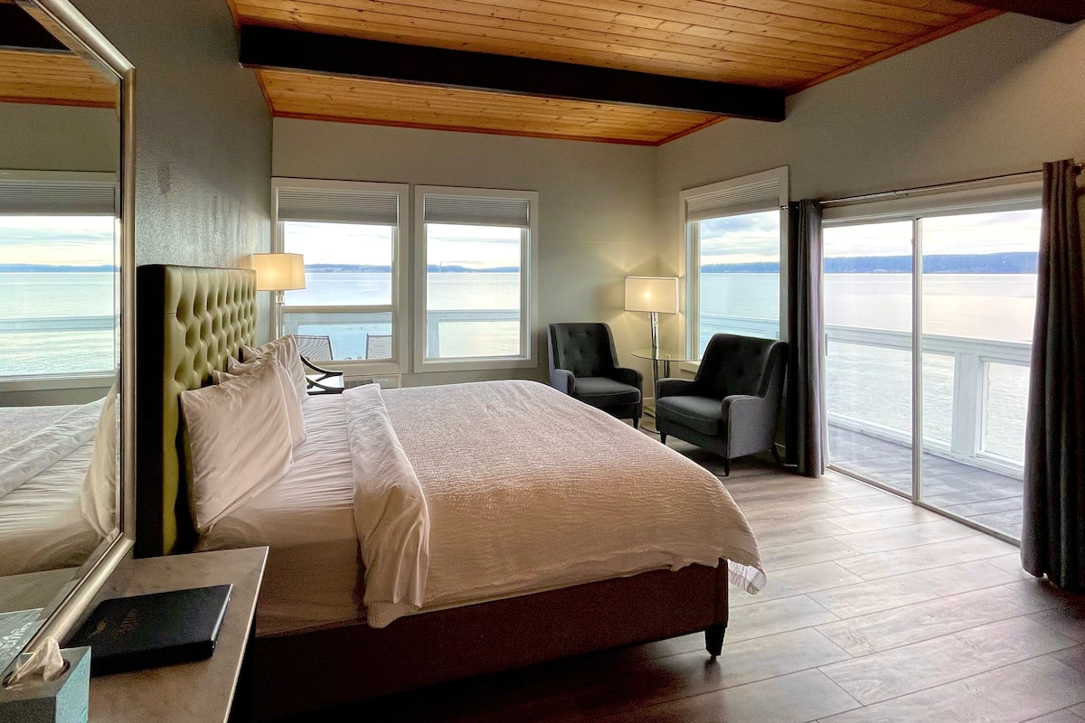 Habitación de hotel con una cama grande, dos sillones, un escritorio y vistas al agua.