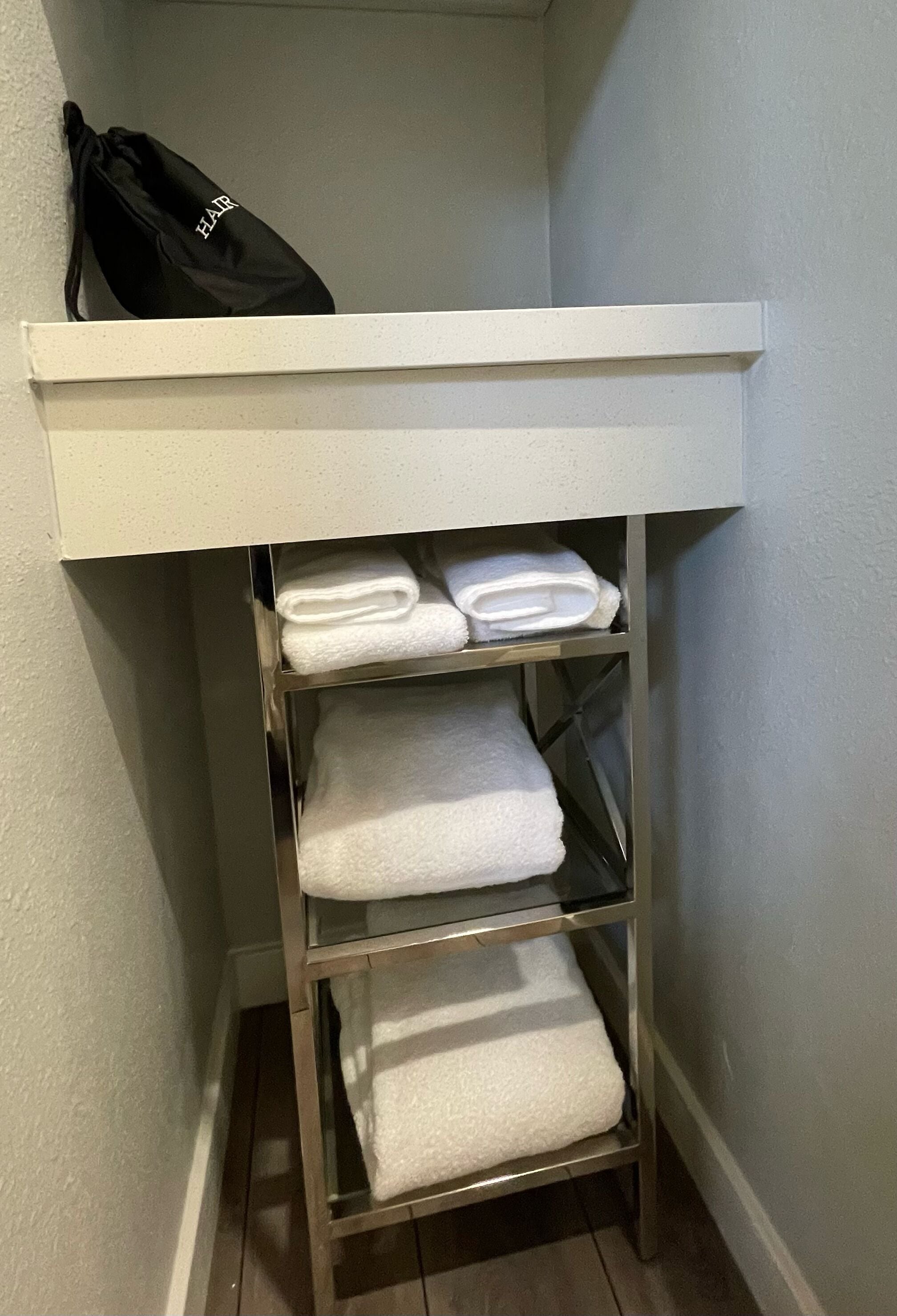 A corner storage unit with a black bag and three shelves holding folded white towels.