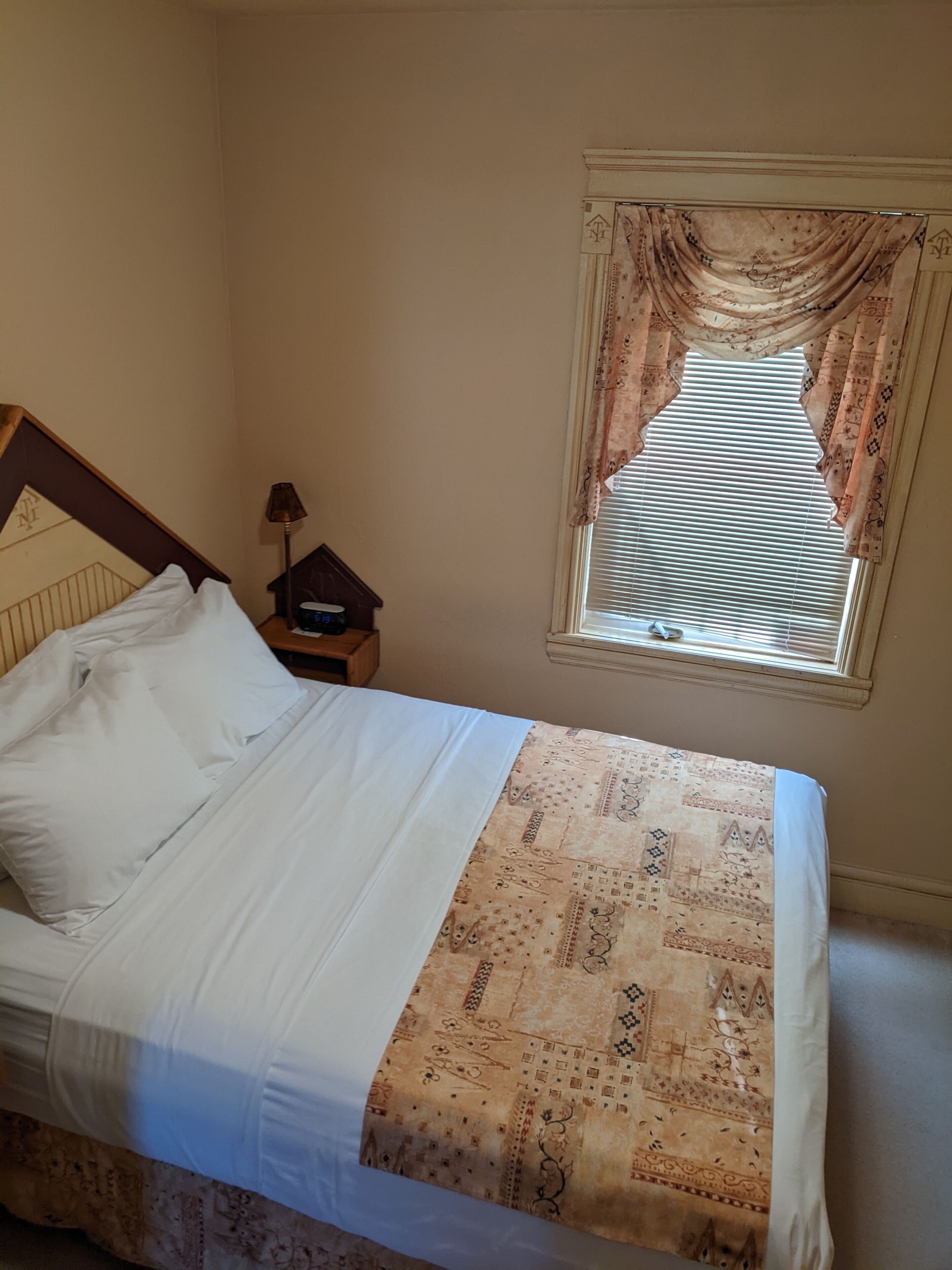 Una cama bien hecha con sábanas blancas y una colcha estampada, una mesita de noche con una lámpara y una ventana con cortinas decoradas.