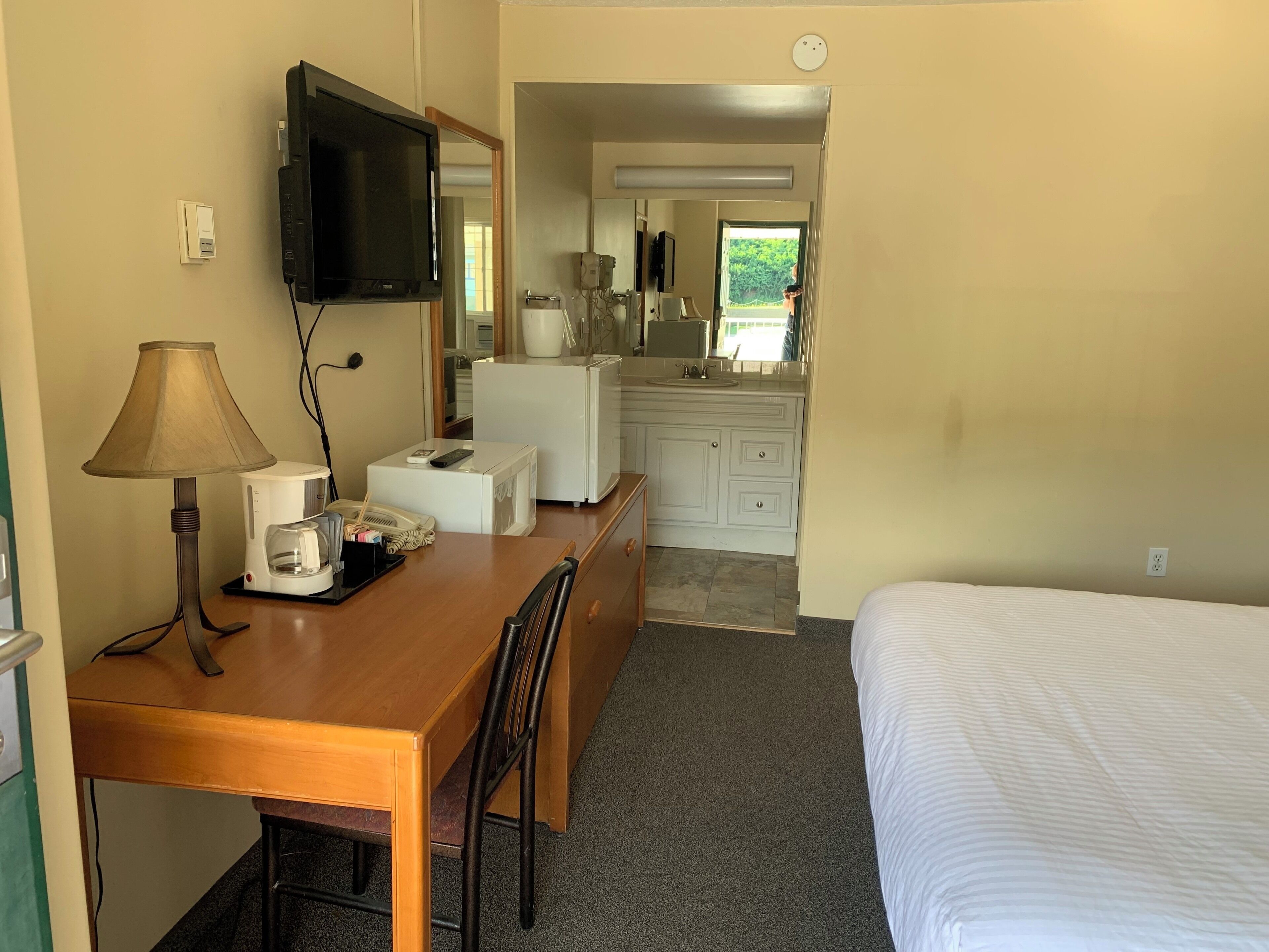 A hotel room with a wooden desk, a lamp, a flat-screen TV, a bed, a bathroom with a sink and mirror, and a window.