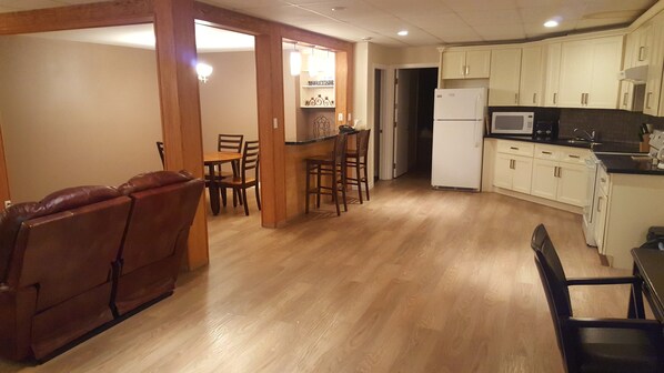 A compact kitchen with a refrigerator, microwave, and wooden cabinets. There is a dining area with a table and chairs, and a leather sofa in the corner.