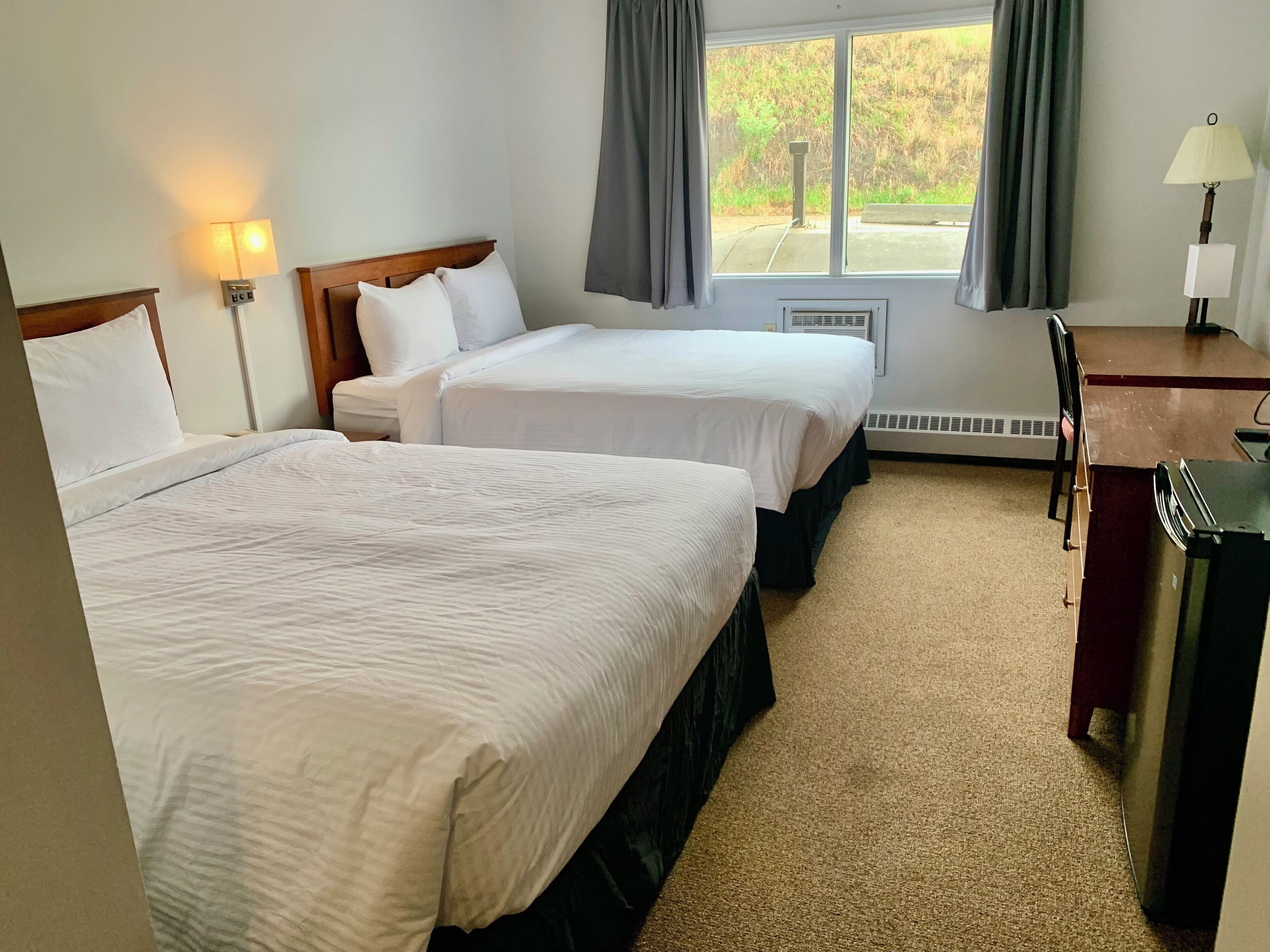 Habitación de hotel con dos camas, un escritorio, una silla, una lámpara y una ventana con cortinas.