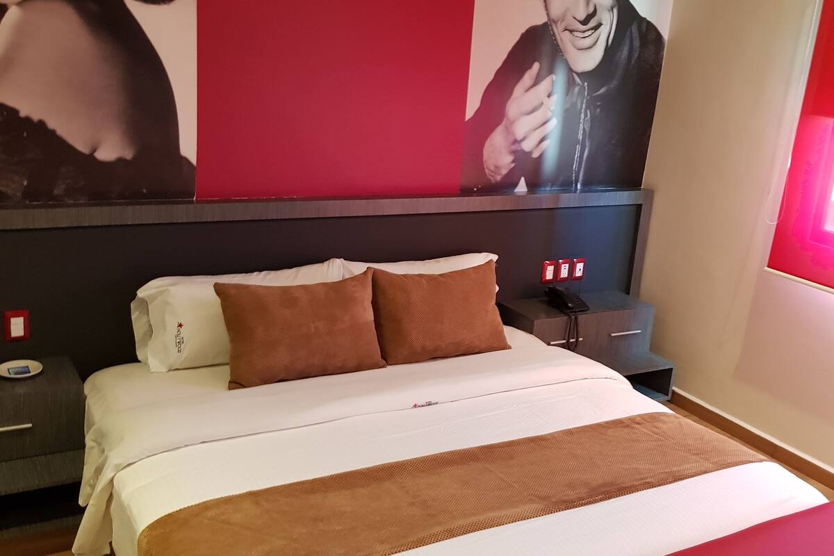 A hotel room with a bed, a nightstand, a red wall, and a large poster.