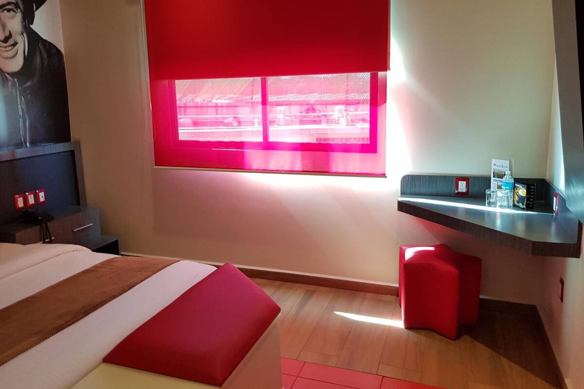 A hotel room with a bed, a desk, a red chair, a window with red blinds, and a wall-mounted TV.