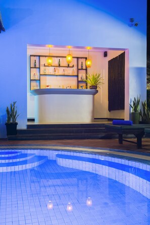 Ein Barbereich am Pool mit Theke, Regalen mit Flaschen, hängenden Lampen und Blumentöpfen.