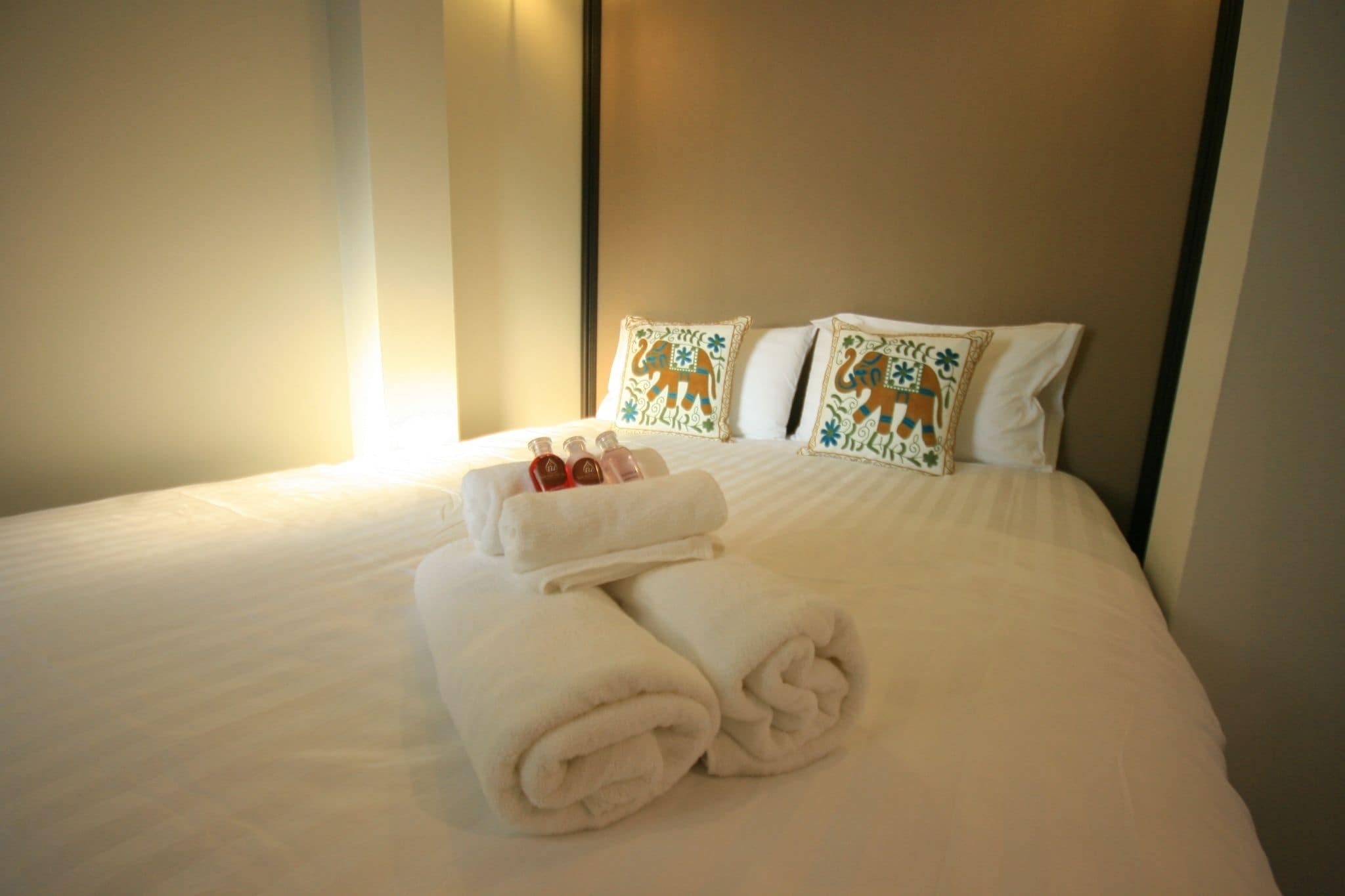 Una cama bien hecha con sábanas blancas, dos almohadas decorativas y un juego de toallas.