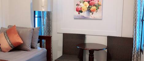 A bedroom with a bed, two wicker chairs, a small round table, a lamp, and a painting of flowers on the wall.