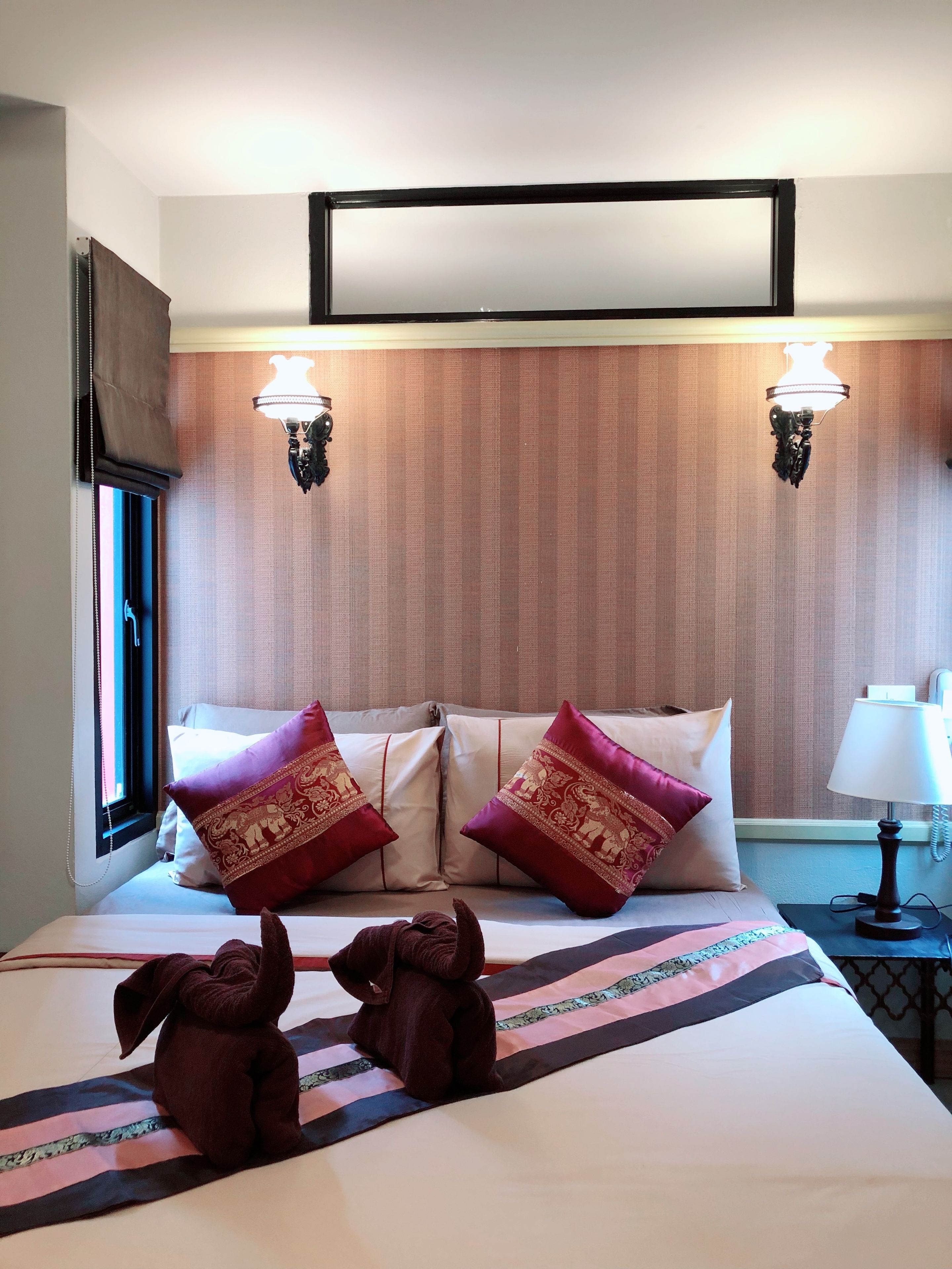 A hotel room with a bed, two decorative pillows, a nightstand with a lamp, and a wall-mounted TV.