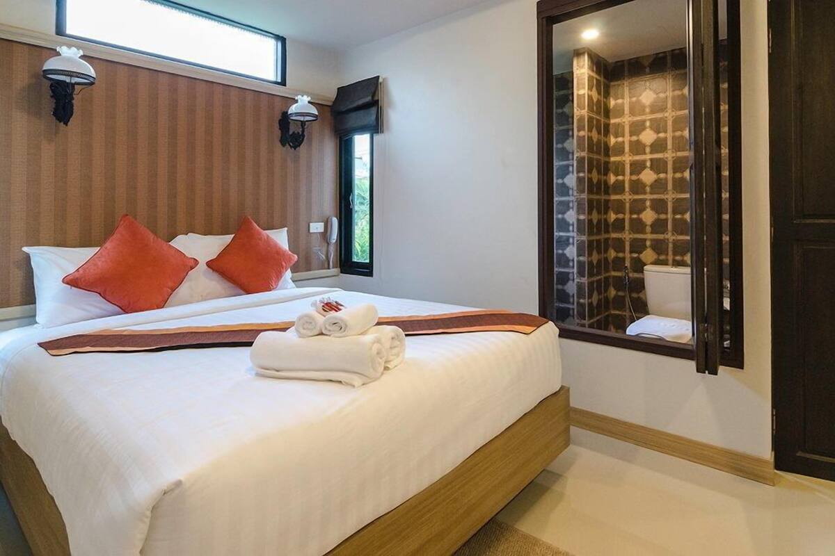 Habitación de hotel con una cama grande, dos almohadas rojas, ropa de cama blanca impecable y un cabecero de madera. Se ve el baño a través de una puerta abierta.
