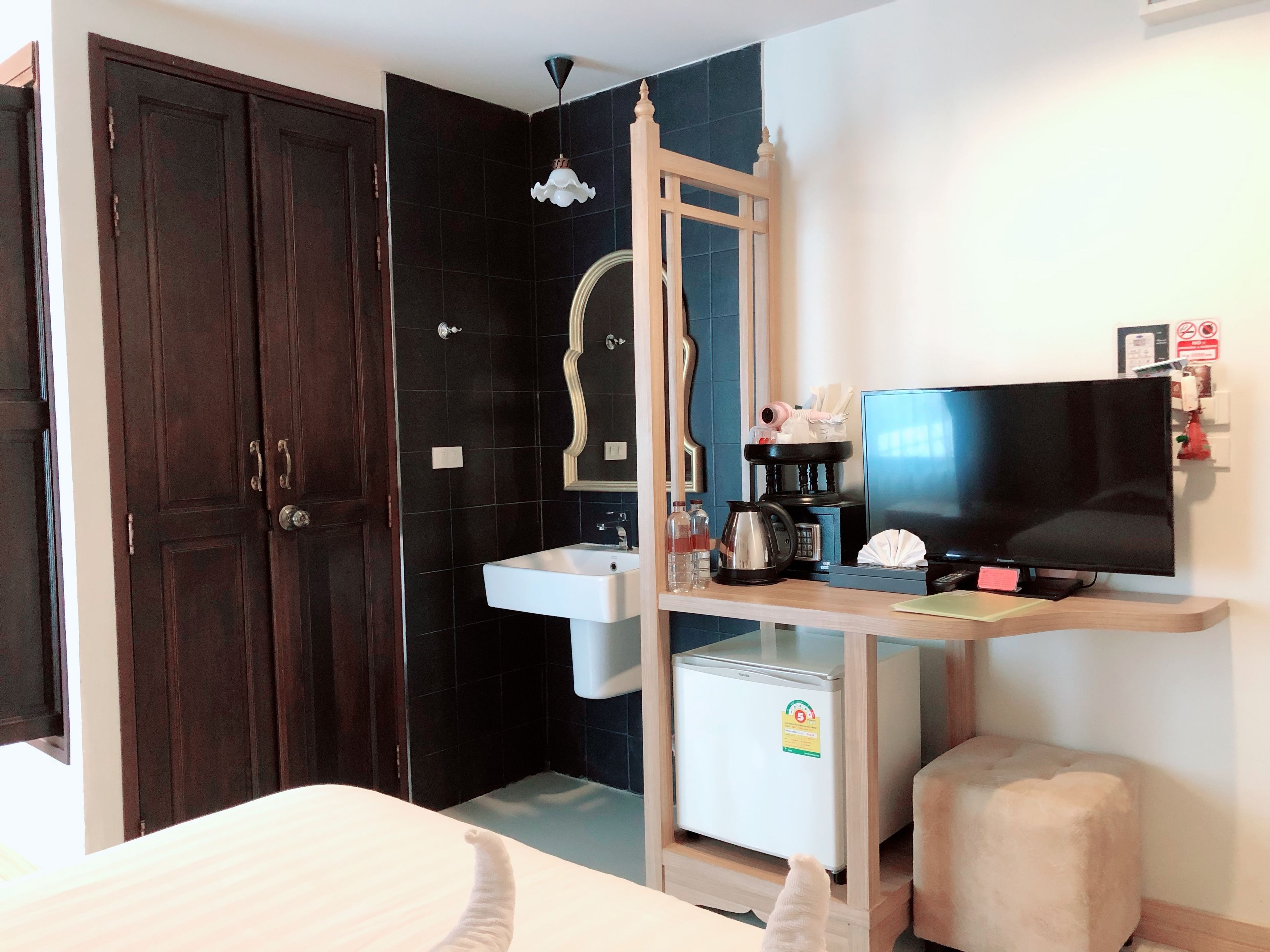 A hotel room with a dark wooden wardrobe, a white sink, a mirror, a television, a small refrigerator, and a bedside table with a lamp.