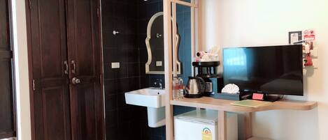 A hotel room with a dark wooden wardrobe, a white sink, a mirror, a television, a small refrigerator, and a bedside table with a lamp.