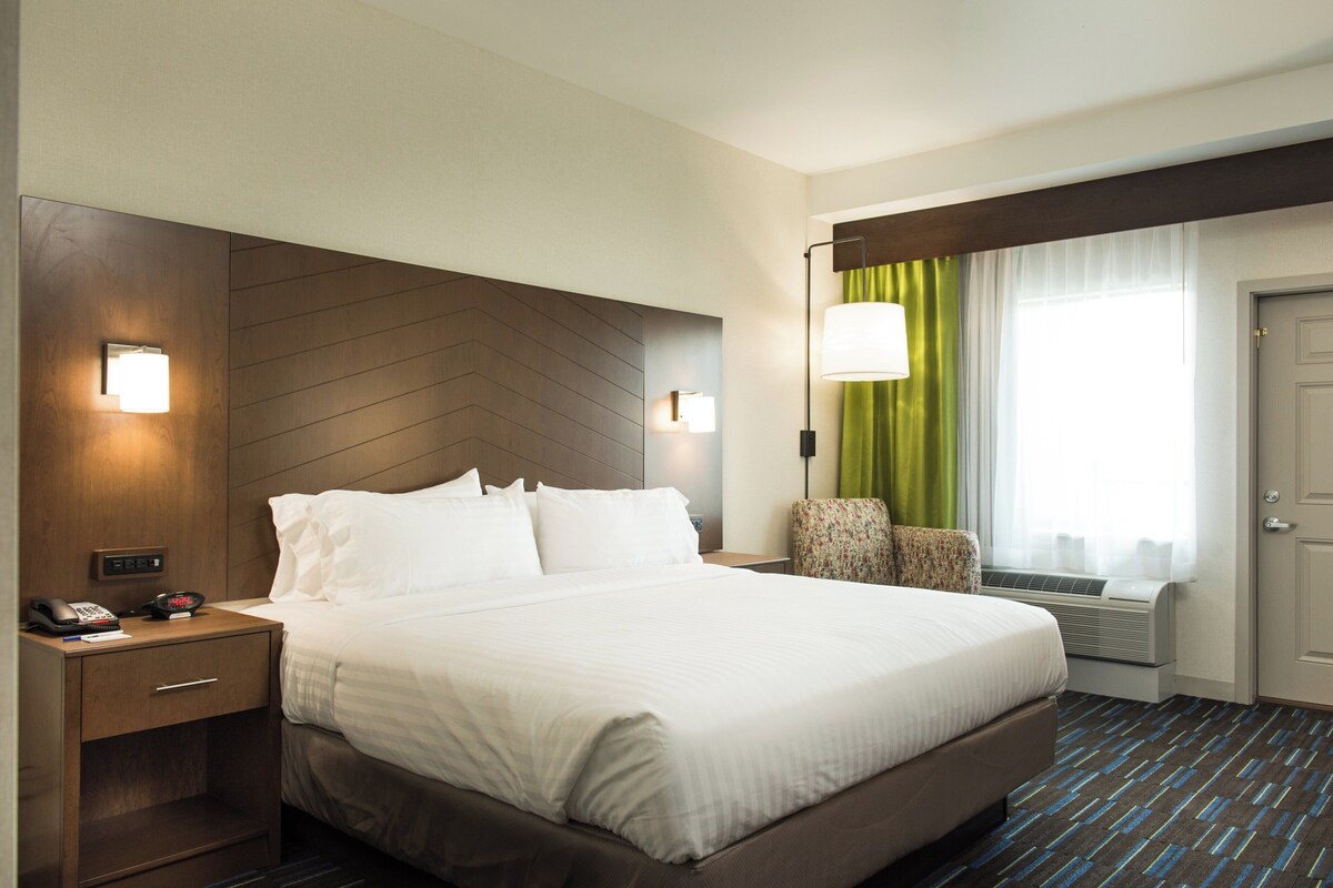 Une chambre d’hôtel avec un grand lit, des tables de chevet, une lampe, une chaise et une fenêtre avec des rideaux.