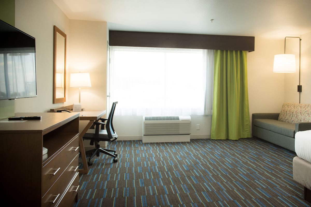 A hotel room with a bed, a desk with a chair, a TV, a window with curtains, an air conditioner, and a lamp.