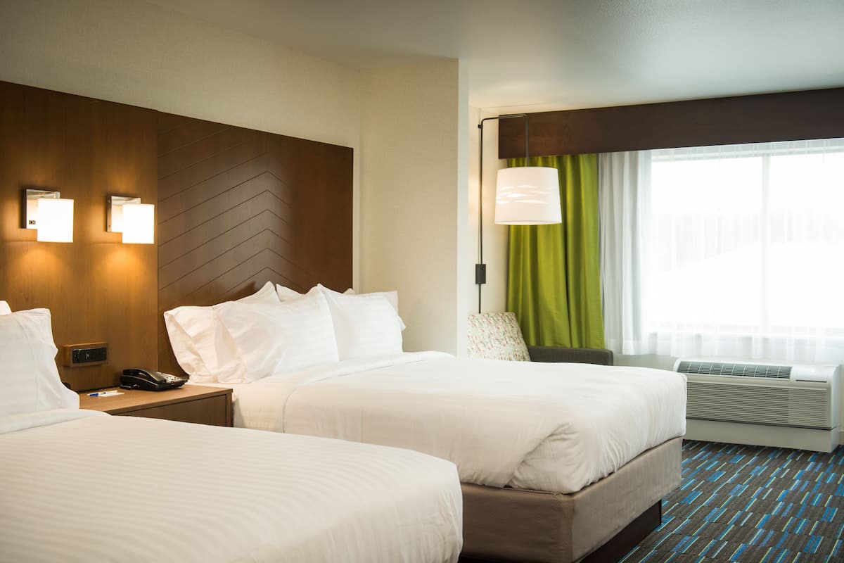 A hotel room with two beds, a nightstand with a phone, a lamp, and a window with curtains.