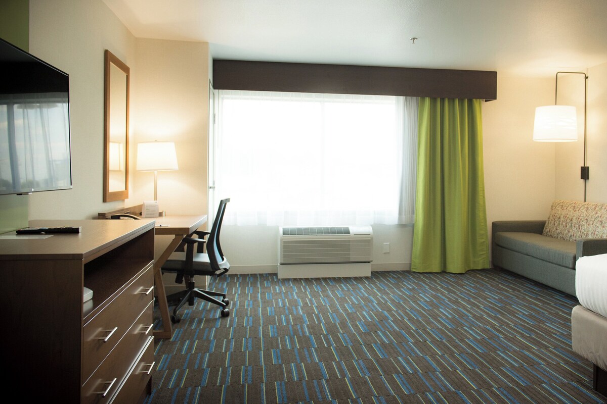 Une chambre d’hôtel comprenant un lit, un bureau avec une chaise, une télévision, une fenêtre avec des rideaux et un appareil de climatisation.