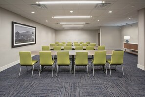 Une salle de conférence avec une longue table et des chaises vertes, un sol recouvert de moquette et un tableau encadré accroché au mur.