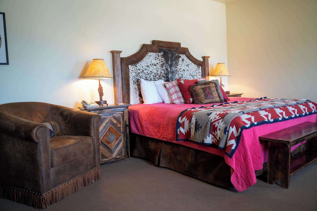 A bedroom with a wooden headboard, a red bedspread, two bedside lamps, a brown armchair, and a patterned rug.